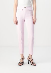 Šviesiai rožinės spalvos pritaikytos kelnės su priekinių kišenėlių, slim fit, ir mažo aukso spalvos metalinio užsegimo detalėmis. Dėvimos su juodais aukštakulniais sandalais su dirželiais.