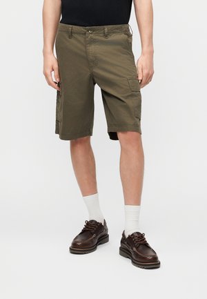 Mężczyzna ubrany w oliwkowe krótkie spodnie cargo, białe skarpetki do kostek, ciemnobrązowe skórzane buty sznurowane oraz czarną koszulkę na jednolitym tle.