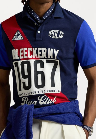 Tengerészkék póló piros átlós csíkkal, fehér grafikával, melyen a "BLEECKER NY 1967" és a "Run Club" felirat található. Kék kötött pulóver a derékon megkötve.