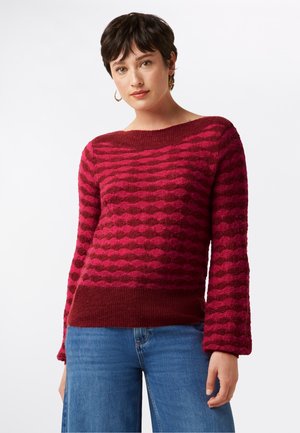 Rosa und burgunderfarbener Pullover mit einem Rautenmuster, strukturierter Oberfläche, lockeren Ärmeln und weitem Ausschnitt, kombiniert mit blauen Jeans.
