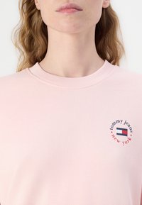 Růžová mikina s ribbovaným kulatým výstřihem, na hrudi s kruhovým logem "tommy jeans" a designem vlajky. Měkká textilie.