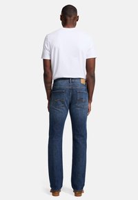 Jeans in denim blu scuro con gamba dritta, due tasche posteriori con cuciture arancioni, indossati con una t-shirt bianca. Patch del marchio discreto in vita.