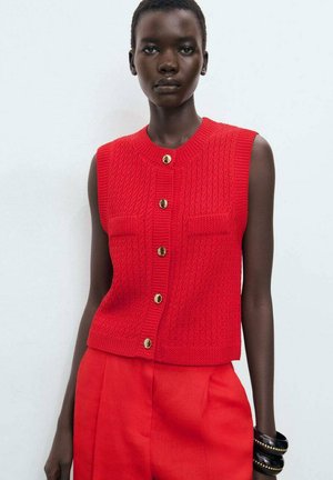 Red knitted sleeveless vest with a round neck, featuring two chest pockets and gold-tone buttons. Worn with matching red trousers.