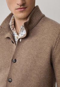 Hnedý pletený kardigán s vysokým golierom, s textúrovanou látkou a tromi čiernymi gombíkmi. Pod ním je vidieť svetlo kockovanú košeľu.