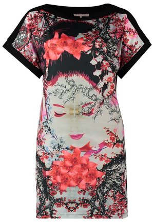 Osoba ubrana w tunikę z krótkim rękawem z nadrukiem czerwonych kwiatów i twarzy gejszy, czarne legginsy i czarne baletki, stojąca na jednej nodze uniesionej do góry.