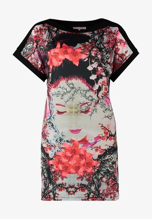 Osoba ubrana w tunikę z krótkim rękawem z nadrukiem czerwonych kwiatów i twarzy gejszy, czarne legginsy i czarne baletki, stojąca na jednej nodze uniesionej do góry.