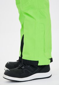 Pantaloni impermeabili verde brillante con rifiniture nere, caratterizzati da un design dritto e polsini regolabili, abbinati a scarpe nere con suole bianche.