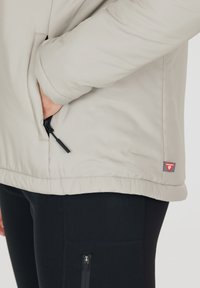 Hand in einer hellbeigen Jackentasche mit schwarzem Reißverschluss, kombiniert mit schwarzen Leggings, die eine Reißverschlusstasche am Oberschenkel haben.