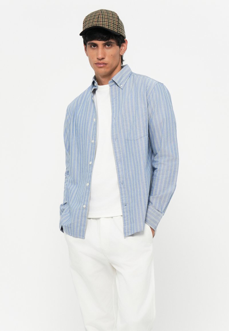 Niebieska koszula w paski z kieszonką, noszona na białym podkoszulku, w połączeniu z jasnymi spodniami i czapką w kratkę.