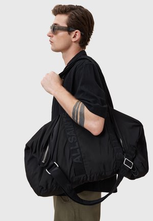 Sac de sport en nylon noir avec une grande forme arrondie, doté d'une fermeture éclair, d'une sangle réglable et d'une marque en relief sur le côté.