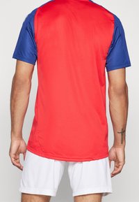 Červený a modrý sportovní dres s krátkým rukávem s hladkým povrchem; nošený s bílými šortkami; viditelný tetování na paži nositele.
