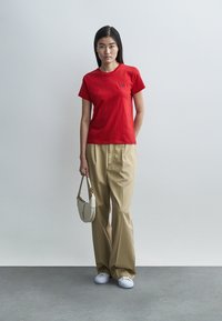 Κόκκινο βαμβακερό μπλουζάκι με στρογγυλή λαιμόκοψη και μικρό λογότυπο, συνδυασμένο με μπεζ παντελόνι φαρδιάς γραμμής και λευκά αθλητικά παπούτσια. Κρατάει μια μικρή λευκή τσάντα.