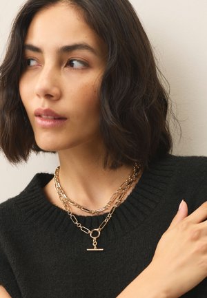 Woman with short wavy dark hair wears layered gold chain necklaces and a black ribbed sweater, looking sideways with a neutral expression.