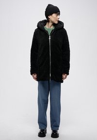 Černá fleecová bunda s texturou, kapucí, zapínáním na zip a dvěma bočními kapsami. Nositelná přes zelený svetr a modré džíny, doplněná černými botami.