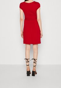 Czerwona sukienka z dopasowanym górnym częściowym i rozkloszowanym dołem, z krótkimi rękawami i zamkiem błyskawicznym z tyłu. W połączeniu z czarnymi sandałami na wysokim obcasie z paskami.