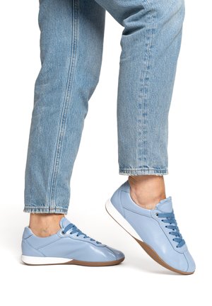 Person wearing light blue leather sneakers with blue laces and light blue jeans, one foot flat and the other slightly raised.