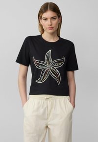 Zwarte cropped t-shirt met een zeesterontwerp in wit, rood en blauwe accenten. De stof lijkt zacht met een ronde hals en korte mouwen.