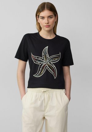 Schwarzes, kurzes T-Shirt mit einem Seestern-Motiv in Weiß, Rot und Blau. Der Stoff wirkt weich und hat einen Rundhalsausschnitt sowie kurze Ärmel.