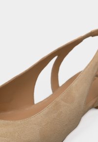 Κοντινό πλάνο μπεζ μοκασίνι από σουέντ με μυτερή μύτη και ανοίγματα στα πλάγια, σε απλό ανοιχτό φόντο.