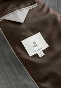 Interior de chaqueta gris entallada, con un forro marrón que tiene un bolsillo en contraste y una etiqueta blanca impresa con "NEXT TAILORED FIT 40R".