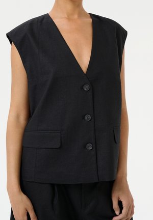 Gilet noir sans manches à boutons, col en V profond et deux poches avant à rabat, porté sur la peau nue.