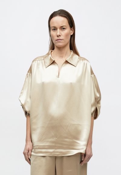 Žena s dlhými hnedými vlasmi oblečená v voľnej, lesklej béžovej saténovej blúzke a zodpovedajúcich nohaviciach, stojaca na jednoduchom bielom pozadí.