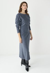 Robe en maille grise à manches longues et silhouette ajustée, dotée d'un ourlet fendu. Associée à des bottes noires à talons hauts.