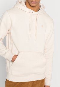 Sweatshirt bege claro com um bolso frontal, capuz ajustável por cordão e uma marcação discreta. Textura suave com punhos e bainha canelados. Design casual.