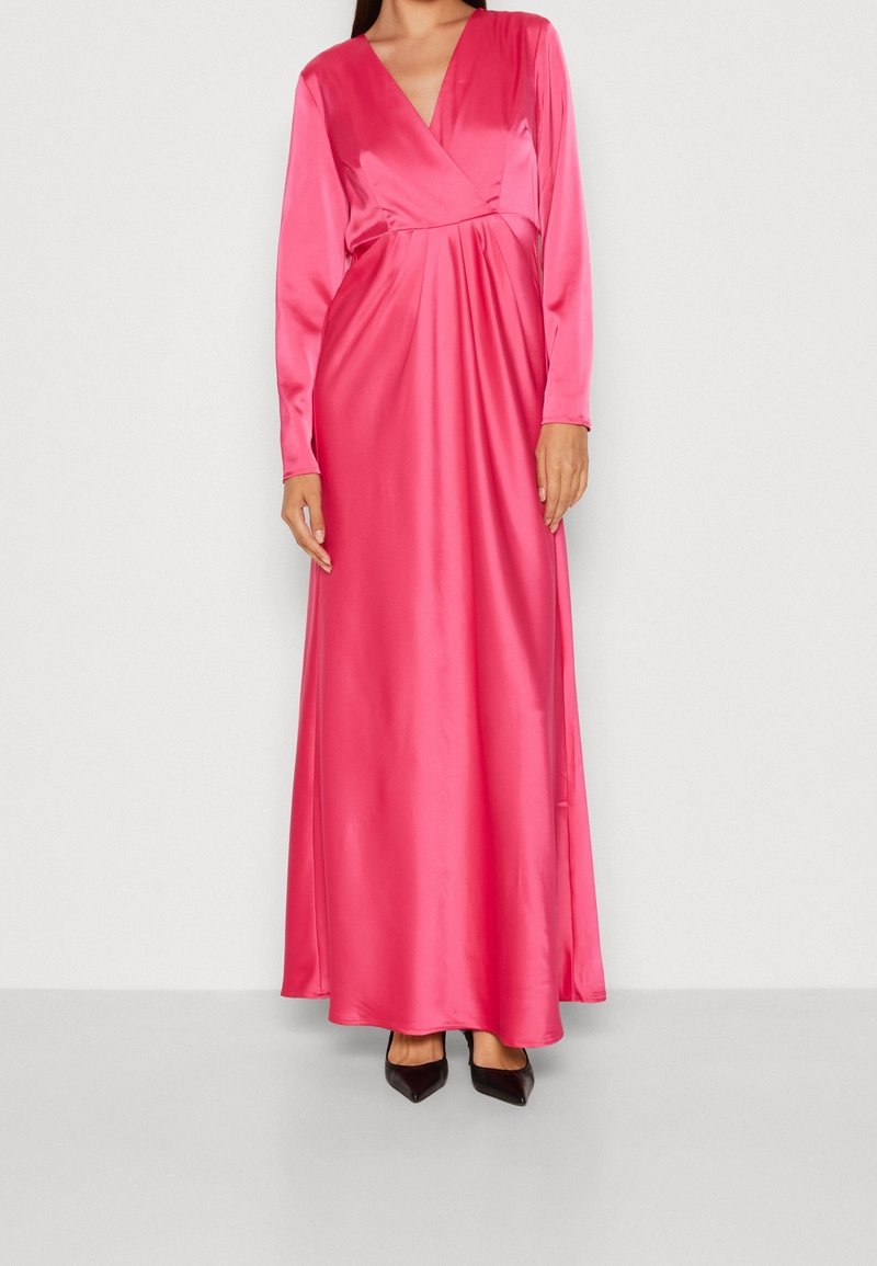 Femme portant une robe portefeuille en satin rose vif à manches longues, associée à des talons noirs à bout pointu, debout devant un fond uni.