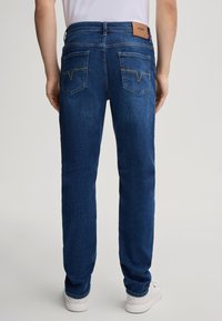 Dark blue denim jeans with a straight cut, featuring two back pockets, light stitching accents, and a brown logo patch on the waistband.