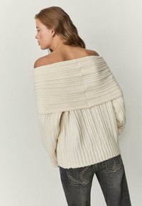 Krem pulover z razkritimi rameni, rebraste teksture in širokim ovratnikom, kombiniran s temnimi kavbojkami z detajlnimi zadnjimi žepi.