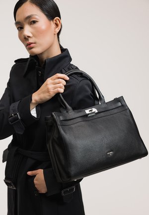 Frau im schwarzen Trenchcoat hält eine strukturierte schwarze Lederhandtasche mit Obergriffen und silbernem Verschluss vor einfarbigem Hintergrund.