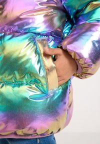 Main dans la poche d'une doudoune brillante et irisée aux teintes violettes, bleues et dorées, contre un fond uni.