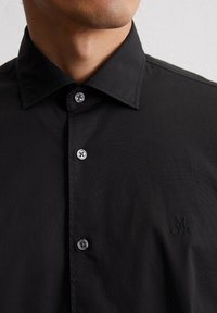 Camicia nera con colletto a punta, dotata di bottoni argentati e logo ricamato sul petto. Tessuto morbido e strutturato.