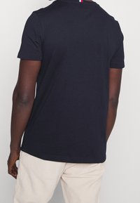 Krátké rukávy, přiléhavé tričko z tmavě modré bavlny. Má klasický kulatý výstřih s barevným akcentem v červené, bílé a modré na zádech.