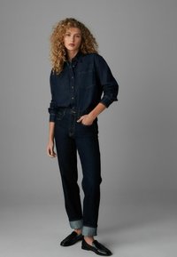 Dunkelblauses Denimhemd mit einer Brusttasche, Knopfverschluss, kombiniert mit dunklen, taillierten Jeans mit umgeschlagenem Saum. Schwarze Slipper runden das Outfit ab.
