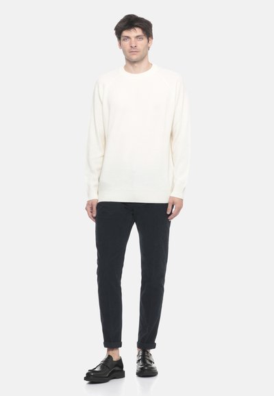 Uomo in piedi che indossa un maglione bianco a maniche lunghe, pantaloni scuri con risvolto e mocassini in pelle nera su uno sfondo bianco semplice.