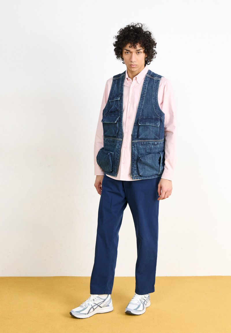 Gilet en denim avec plusieurs poches, porté sur une chemise boutonnée rose pâle, associé à un pantalon bleu et des baskets de couleur claire.