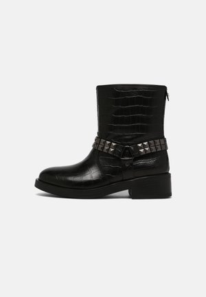 Botte cheville en cuir noir embossé crocodile avec talon épais et bride cloutée décorative. Fermeture éclair latérale pour un enfilage facile. Surface texturée.
