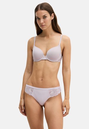 Mujer con cabello largo y castaño, vestida con un sujetador lila claro y bragas a juego con encaje, de pie sobre un fondo blanco liso.