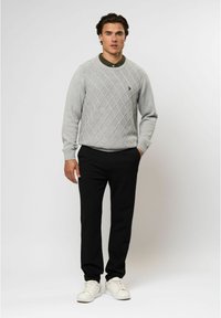 Pilkas tekstūruotas megztinis su deimanto raštu, juodos kelnės ir baltos sportbačiai. Modelis stovi atsukęs į priekį, rankos kišenėse.