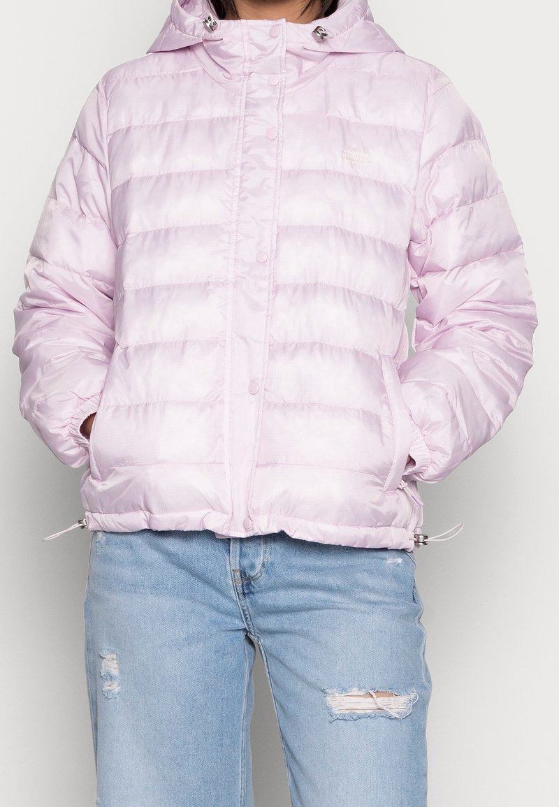 Giacca puffer rosa chiaro con cappuccio, caratterizzata da un design trapuntato e chiusure a scatto, abbinata a jeans blu strappati.