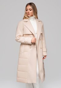 Žena v béžovém dlouhém prošívaném a vlněném kabátu přes bílý rolák, dívající se dolů a držící límec kabátu jednou rukou.