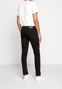 Osoba stojí zády k fotoaparátu, má na sobě bílé tričko a černé slim-fit džíny, doplněné o bílé tenisky.