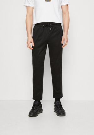 Mężczyzna w czarnych spodniach z sznurkiem, czarnych tenisówkach i białej koszulce, stojący na tle jednolitego białego tła.