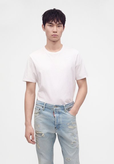 Jeune homme aux cheveux noirs courts, portant un t-shirt blanc uni et un jean déchiré bleu clair, debout avec une main dans la poche.