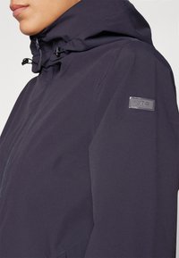 Marineblå vanntett jakke med hette, med glidelås foran, justerbare snorer og en logo-patch på overarmen. Glatt tekstur.
