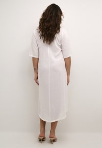 Robe en lin blanche avec des manches courtes, longueur mi-mollet et un détail de couture centrale. Portée avec des talons légers et ouverts.