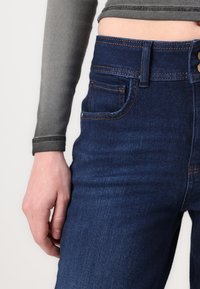 Tamno plavi traperice s narančastim šavovima, s pet džepova i prednjim zatvaranjem na dugme. Glatka tekstura i slim-fit dizajn.