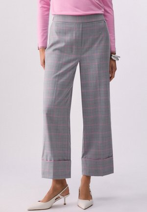 Persona che indossa pantaloni grigi a quadri a gamba larga con linee rosa, una maglia rosa a maniche lunghe, scarpe bianche con tacco e un bracciale d’argento.